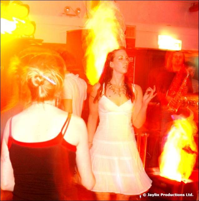A woman in a white dress dances beside large flames, with a person in a red top in front