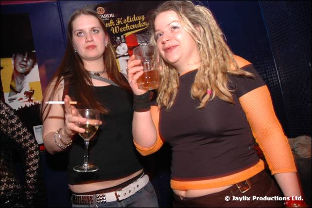 Two women pose holding drinks, one with a cigarette, in front of a poster