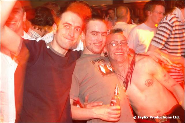 Three men pose together with arms around each other, the one on the right shirtless and wearing glasses