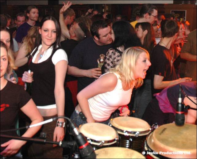 A woman in a white top leans over conga drums with microphones, in a crowd