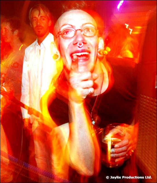 A person with glasses and a nose ring sings or shouts into a microphone while holding a drink