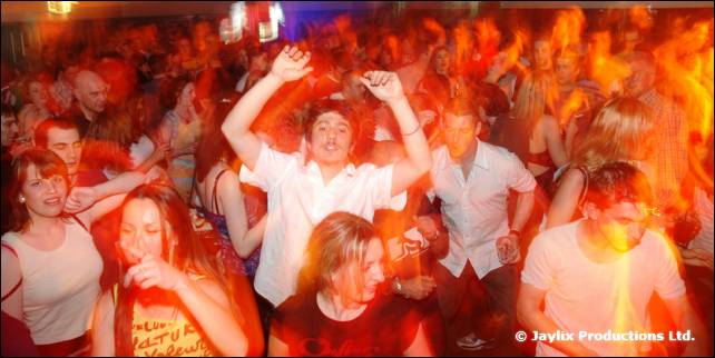 A packed crowd dances with arms raised in red and orange light