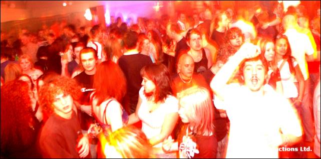 A large crowd dances in a hall, a person on the right with arms raised