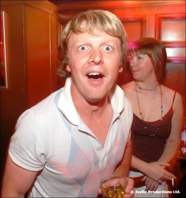 A man with blond hair and wide eyes holds a drink, with a woman behind him
