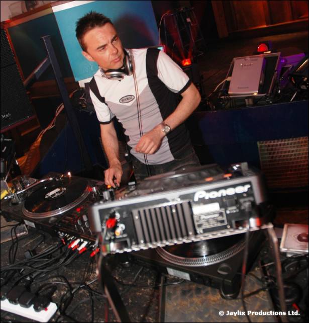 A man in a black and white top with headphones stands at Pioneer DJ decks