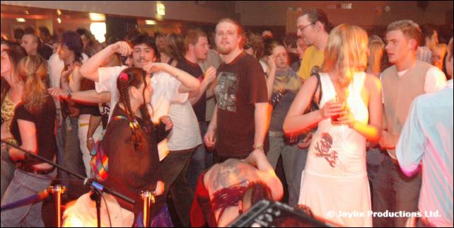 A crowd dances in a large room, a woman in a white dress at the centre right