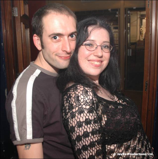 A man and a woman in glasses and black lace pose together in a doorway