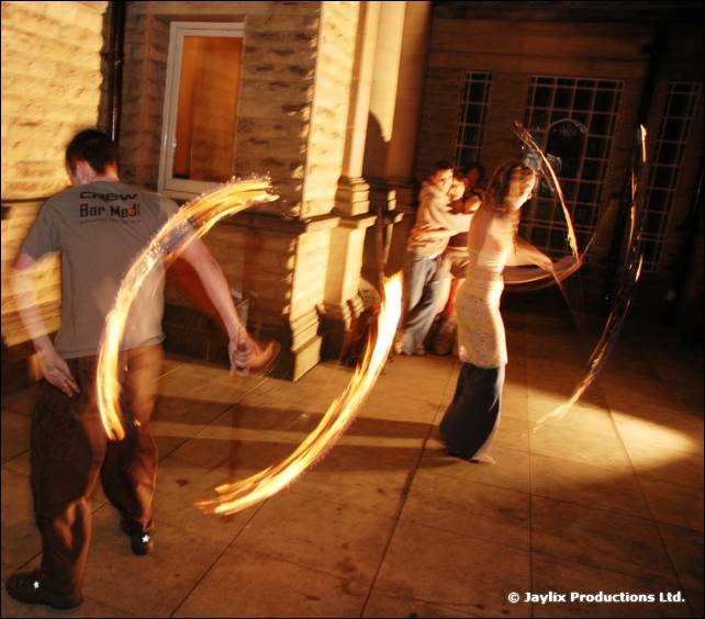 Two people spin fire poi outside a stone building at night, the nearer one in a Bar Med shirt