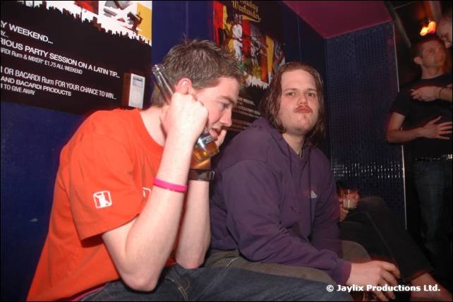 Two men sit close together, one holding a bottle to his face, in front of posters