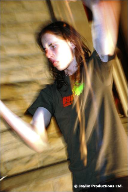 A person with long hair raises an arm, blurred with motion in front of a stone wall