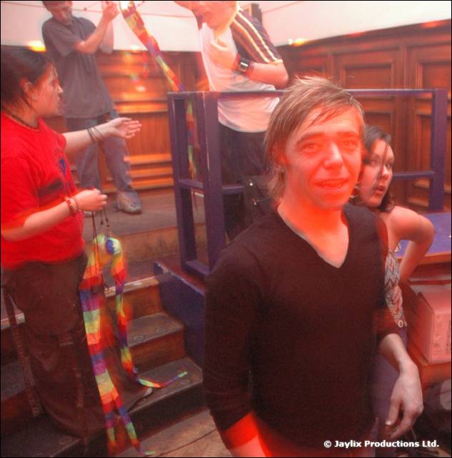A man in a black v neck top looks at the camera beside a wooden stage with people behind