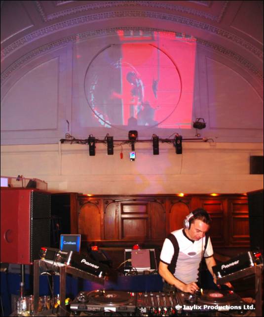 A DJ in a white top with braces stands at Pioneer decks in a hall with a red projection behind