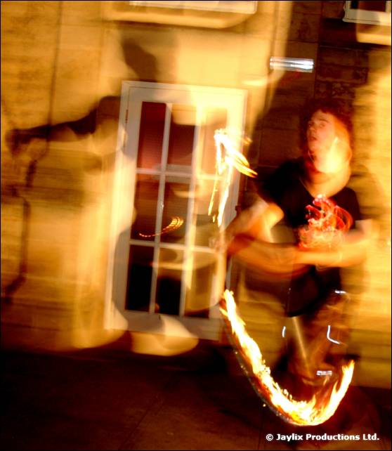 A person in black swings fire beside a lit window on a stone wall at night