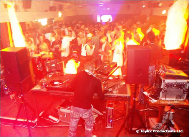 A DJ stands behind decks and speakers facing a crowd, with flames on either side