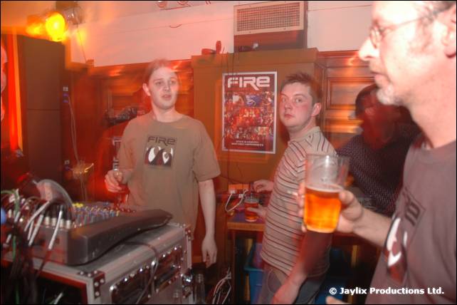 Men stand behind a mixer and DJ equipment, one holding a pint, with a FIRE poster behind