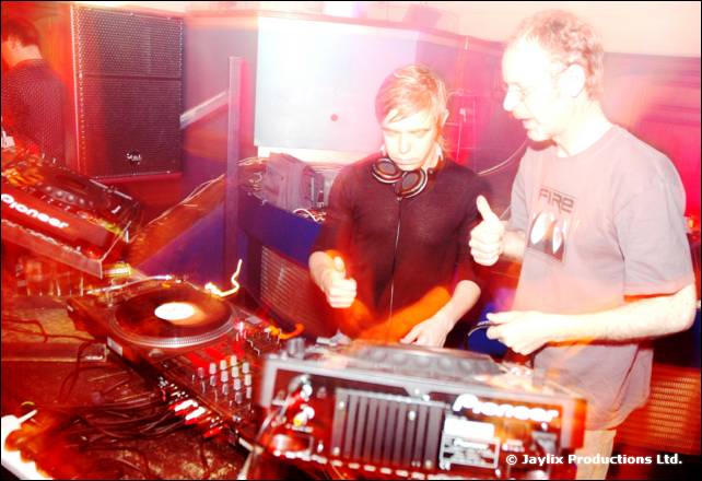 Two men in headphones and a FIRE t-shirt at the decks, giving thumbs up