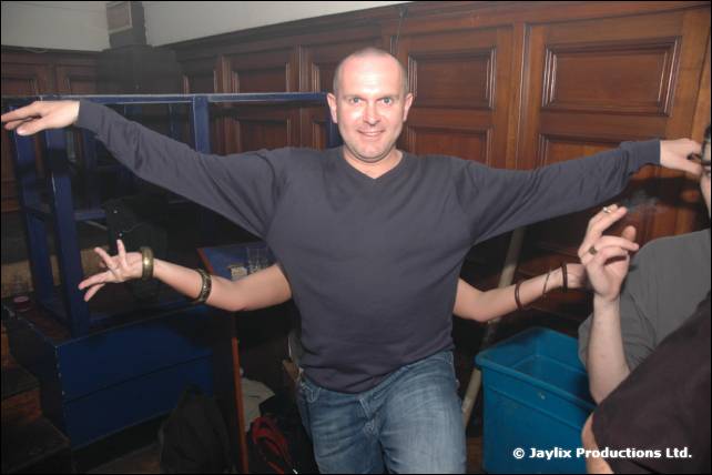 A man in a dark jumper stands with arms outstretched in front of a blue frame