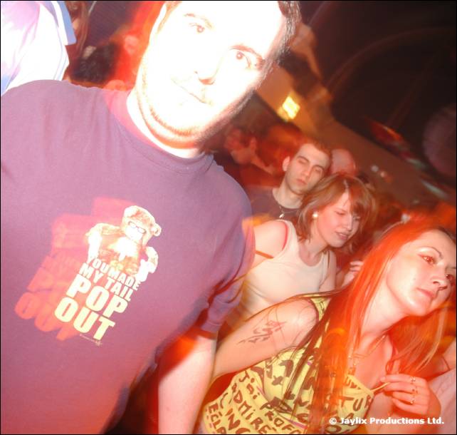 A man in a dark t-shirt leans into the camera, with dancing people behind him