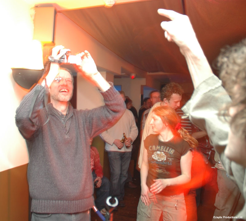 A man in a grey jumper holds a camera up above the crowd while a woman in a dark top dances behind him