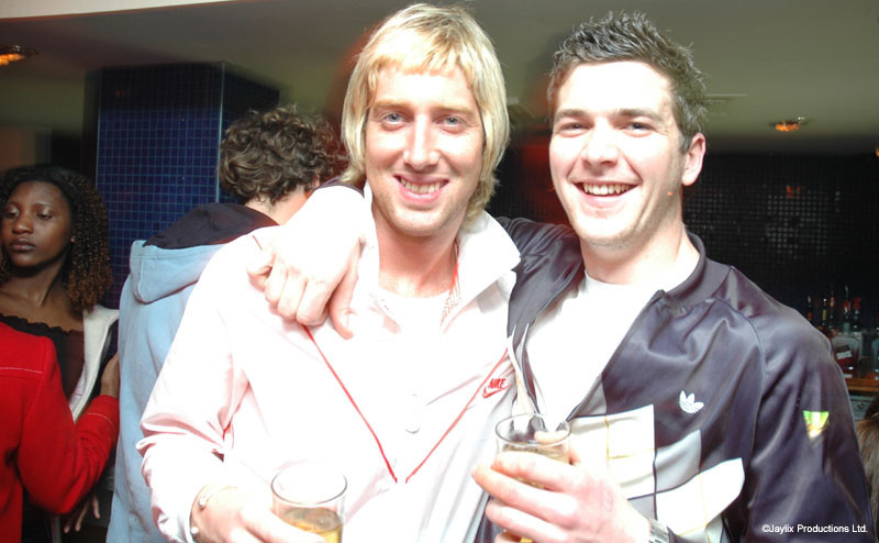 Two smiling men in light tracksuit tops hold drinks with their arms around each other