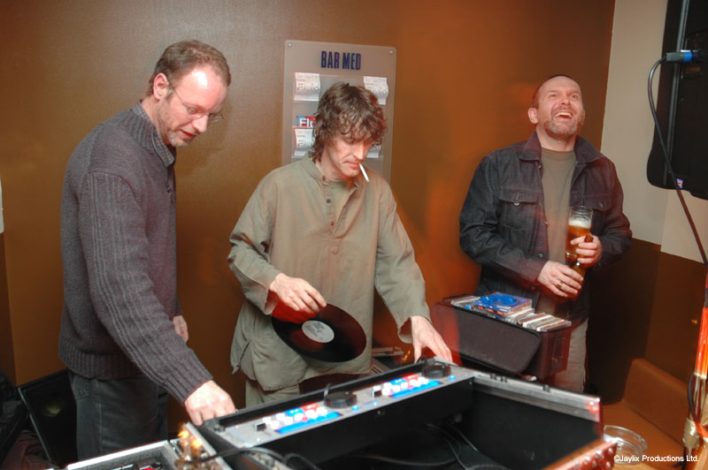 Three men at DJ decks, the one in the middle holding a record, a Bar Med sign on the wall behind