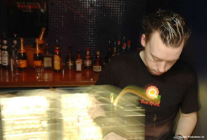 A bartender with spiky hair in a black Bar Med t-shirt works at a bar with bottles behind