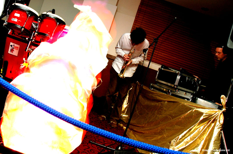 A musician plays a saxophone near red congas, with a large glowing fire prop beside a blue rope