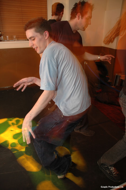A man in a grey t-shirt dances with his knees bent on a dark floor with green and yellow lights