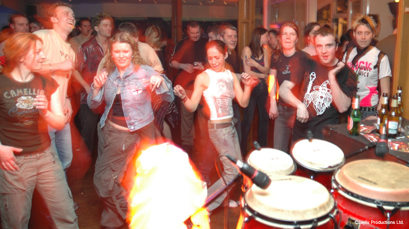 A crowd dances around red congas in a bar, with a glowing fire prop on the floor