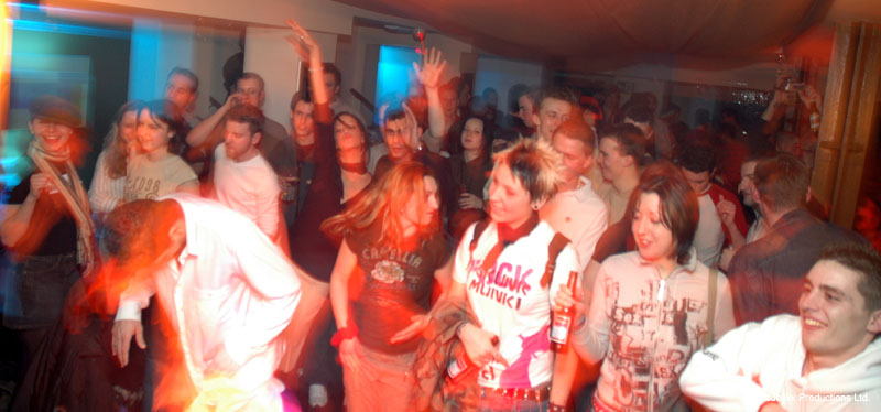 A crowd dances in a busy bar, many in matching white and grey t-shirts