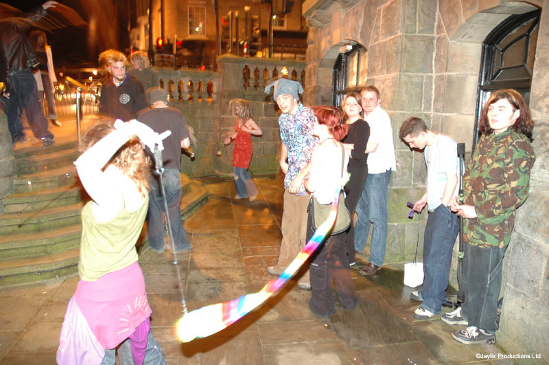 A person swings a staff with a rainbow cloth while onlookers stand on stone steps at night