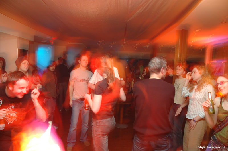 A busy dance floor in a bar under a draped ceiling, lit with orange and pink