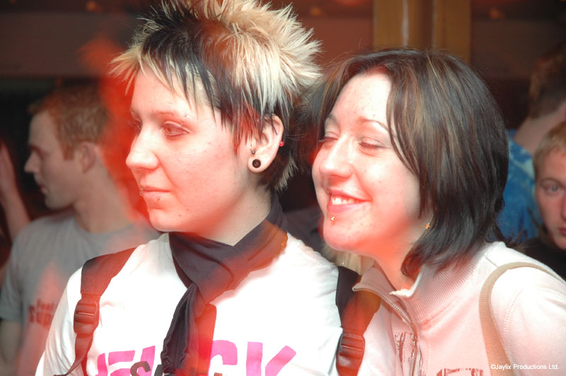 Two women smile and look to the side, one with spiky hair and a black scarf, in white tops