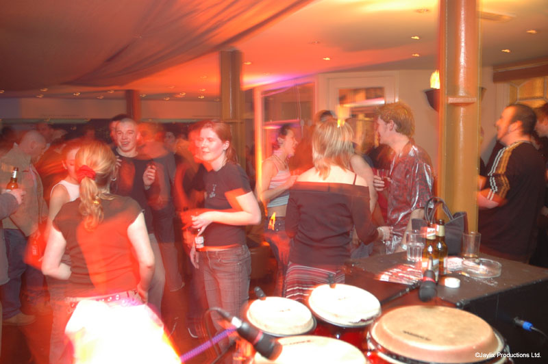 A crowd in a bar, with a woman in a red top from behind and congas on a table on the right