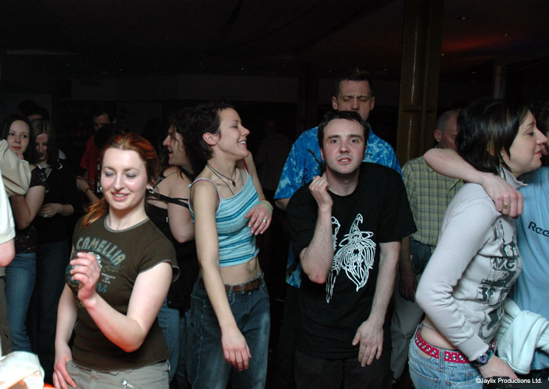 A group of people stand and dance in a dark bar, looking towards the camera