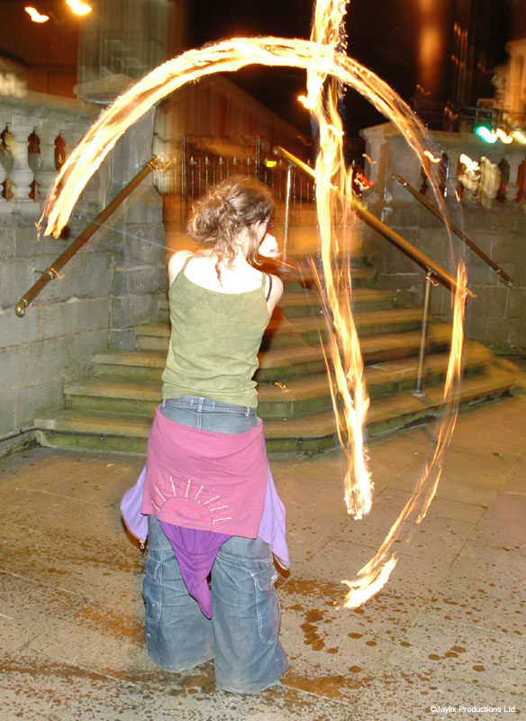 A person in a green top and pink jumper tied at the waist spins fire poi on stone steps at night