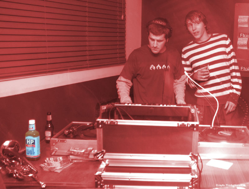 Two people stand behind DJ equipment in red-toned light, with a bottle of HP sauce and a trumpet on the table