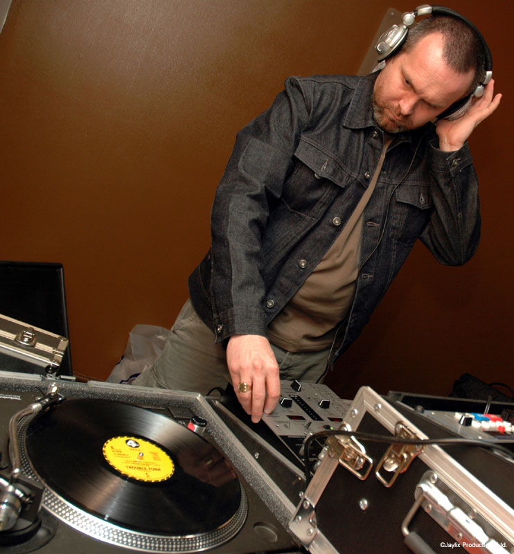 A DJ in a denim jacket and headphones leans over a turntable and mixer