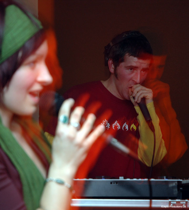A woman in a green headscarf sings in the foreground while a man in a red t-shirt performs behind her