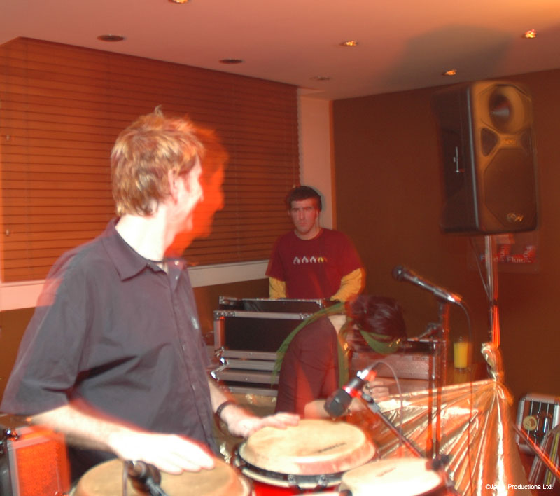 A man in a dark shirt plays congas while a person stands behind DJ equipment and a speaker