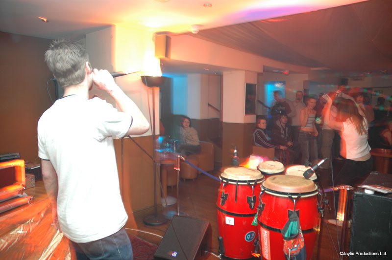 A person in a white T-shirt sings into a microphone beside red conga drums in a bar