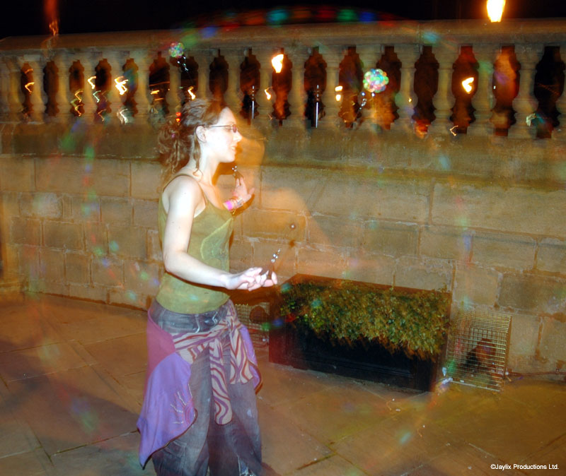 A person in a green top and patterned skirt stands on a stone terrace with fire poi blurred behind