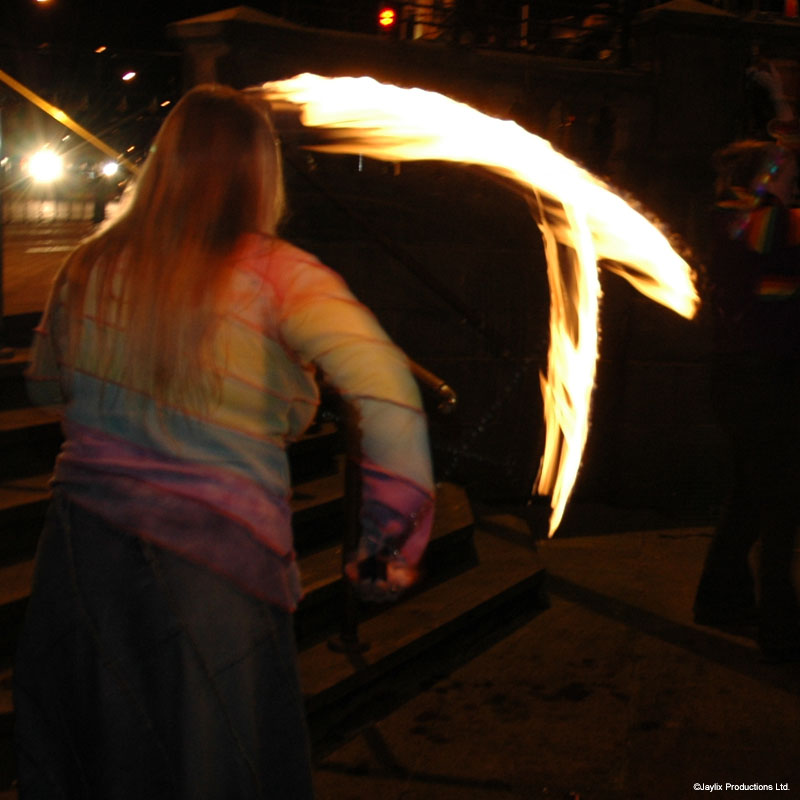 A person with long hair swings a flaming staff in an arc at night, seen from behind