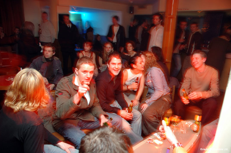 A group sits around a table in a busy bar, one person pointing at the camera