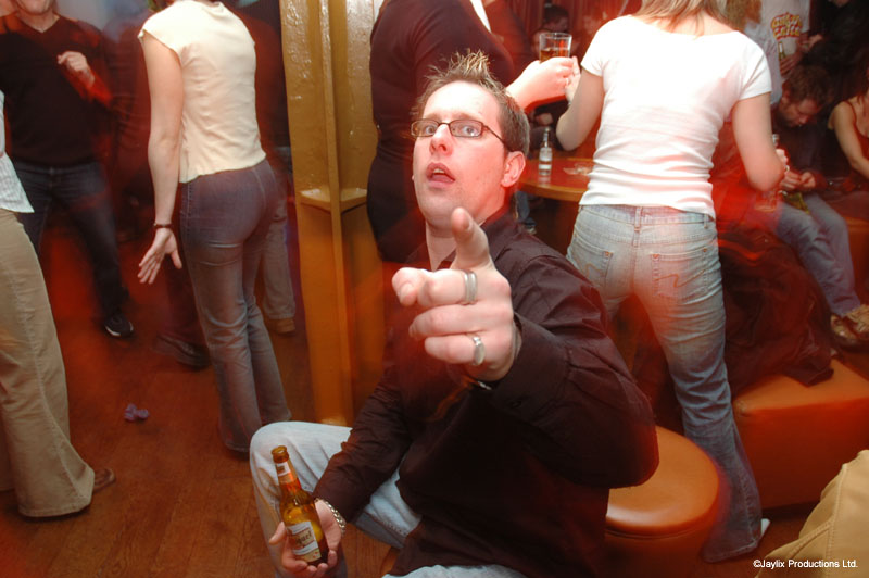 A man with glasses points at the camera in a crowded bar, holding a bottle