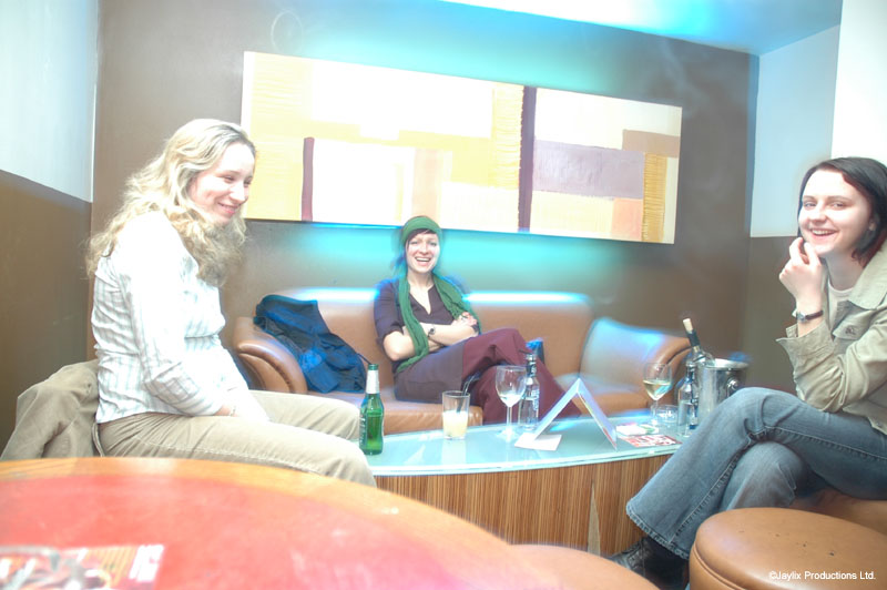 Three people sit and talk on sofas around a low table with bottles and glasses