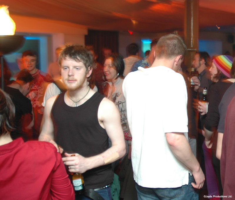 Two men stand in a crowded bar, one in a black vest and one in a white T-shirt
