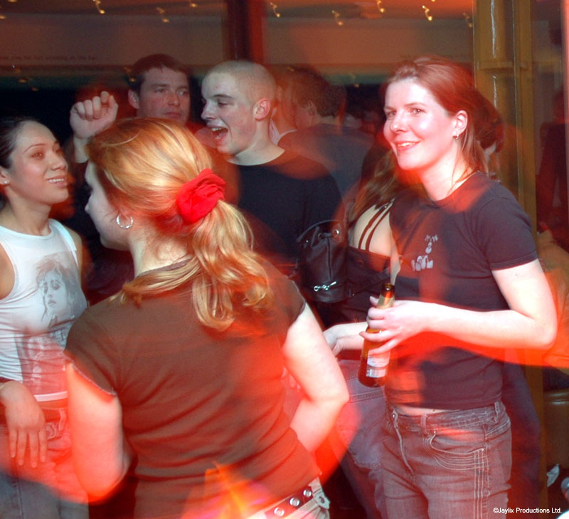 A crowd dances in a bar, a woman in a dark T-shirt smiling and holding a bottle