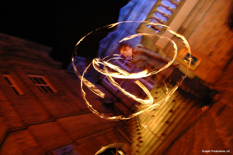 Fire poi trails swirl at night in front of a stately building, with a blurred figure behind