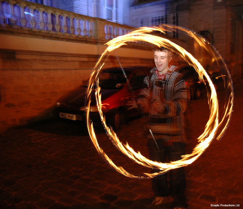 A person in a striped hooded top spins two fire hoops outdoors at night beside a parked car